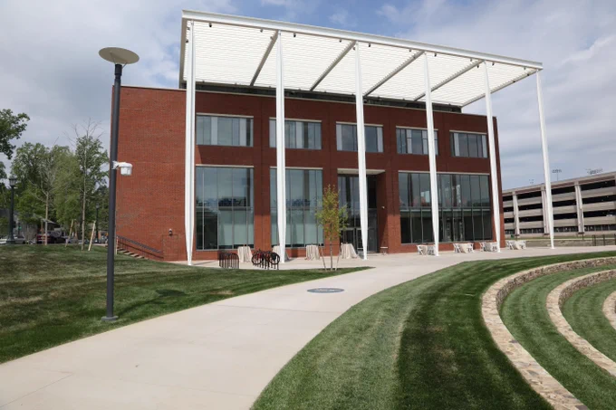 The School of Data Science building, featuring large windows and a prominent white awning supported by steel beams, fronted by a stunning lawn with a curved pathway. In the background, a multi-level parking garage is visible. The setting suggests a campus-like environment, emphasizing open spaces and contemporary architecture.