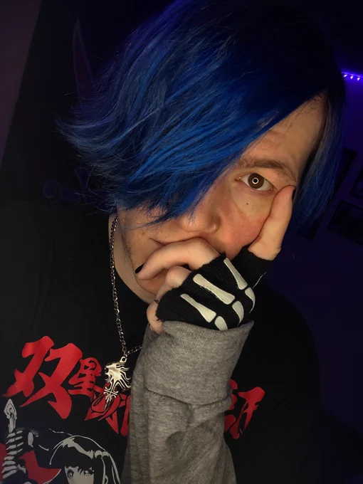 Local Twitter/YouTube/TikTok sensation Super Nerd Daniel, posing for a selfie in front of their cell phone's camera at a somewhat-tilted angle, with his left hand positioned against his face, the index finger raised to rest against their cheek.

Daniel is a white nonbinary person with dyed blue hair and hazel-brown eyes, wearing a black t-shirt with red and white graphics on it over a long-sleeved light grey shirt, black fingerless gloves with white skeleton hand patterns, and a small silver necklace modeled after that of Squall Leonhart from the video game Final Fantasy VIII.