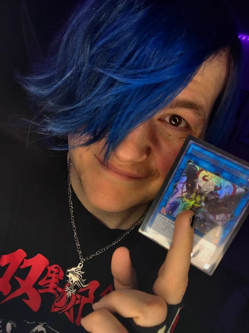 Local Twitter/YouTube/TikTok sensation Super Nerd Daniel, posing for a selfie in front of their cell phone's camera at a somewhat-tilted angle, holding a copy of the Yu-Gi-Oh TCG Link Monster Card "Condemned Darklord" in between two fingers of his left hand.

Daniel is a white nonbinary person with dyed blue hair and hazel-brown eyes, wearing a black t-shirt with red and white graphics on it over a long-sleeved light grey shirt, black fingerless gloves with white skeleton hand patterns, and a small silver necklace modeled after that of Squall Leonhart from the video game Final Fantasy VIII.