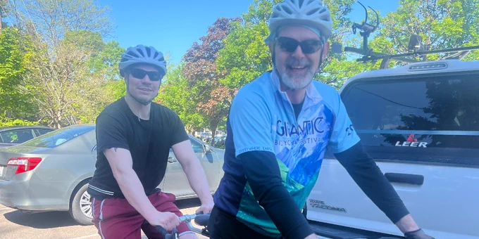 Pictured is Athlete Adam and Volunteer John on a tandem bike, smiling at the camera. They are both geared up in the parking lot near the group and ready to hit the trail.