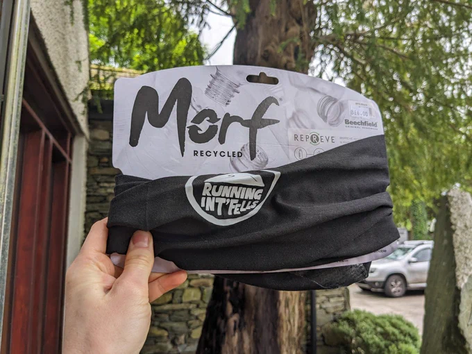 A hand holding up a black Beechfield Morf running buff in its cardboard packaging, with the white Running Int Fells logo on it