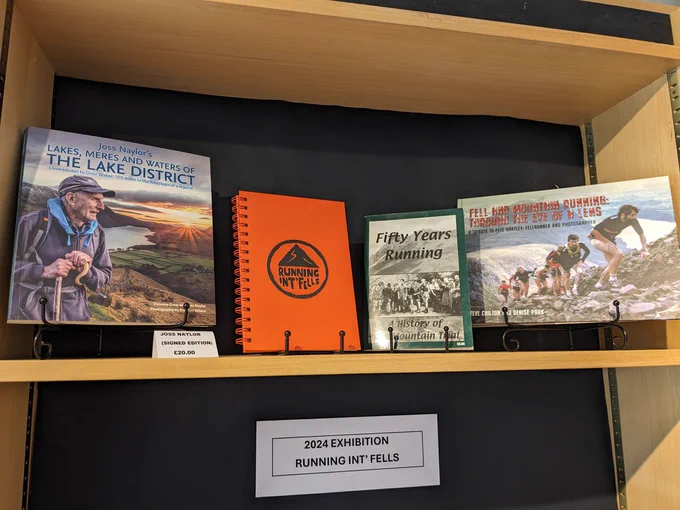 A selection of three books and an orange notebook with the black Running Int Fells logo on the front, displayed on a book shelf in The Armitt shop. On the left is Joss Naylor's Lakes, Meres and Waters of the Lake District. In the middle, right of the orange notebook, is Fifty Years Running by the Fell Runners' Association. On the right is Fell and Mountain Running Through the Eye of the Lens by Steve Chilton and Denise Park