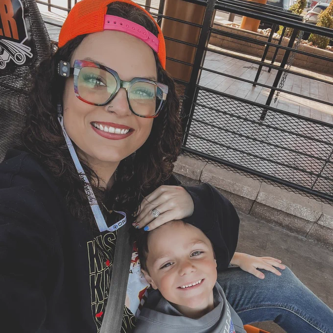 Ashley Mizell and her young son enjoy a ride at an amusement park. Ashley, wearing the OrCam MyEye and a bright orange hat, smiles alongside her son.
