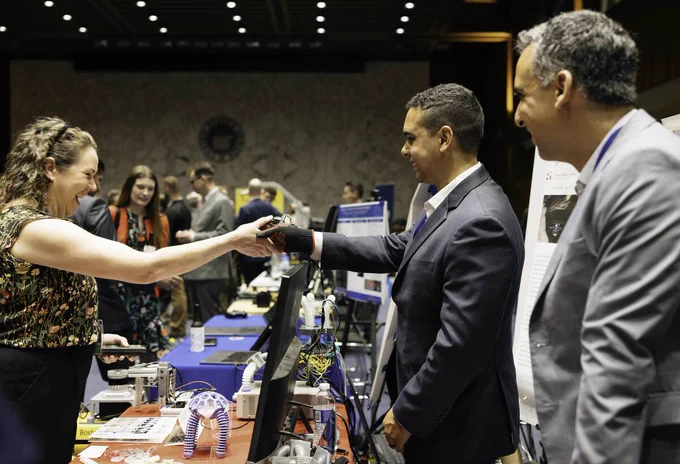 A Congressional staffer shakes hands with graduate student Richard Desatnik. Desatnik is wearing a glove that remotely operates a soft robot, on table below. 
