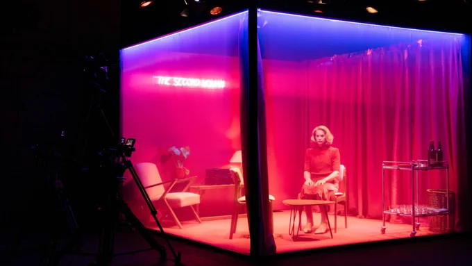 A person sits in a theatre set of a room with a few chairs, a coffee table and a drink trolley. The room is bathed in pink light and there is a neon sign on the wall that reads ‘The Second Woman’.
