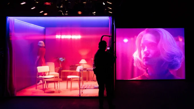 A person stands in a room bathed in pink light facing a wall. A videographer stands in front of the set with their camera. To the right, a screen shows the actor’s face.
