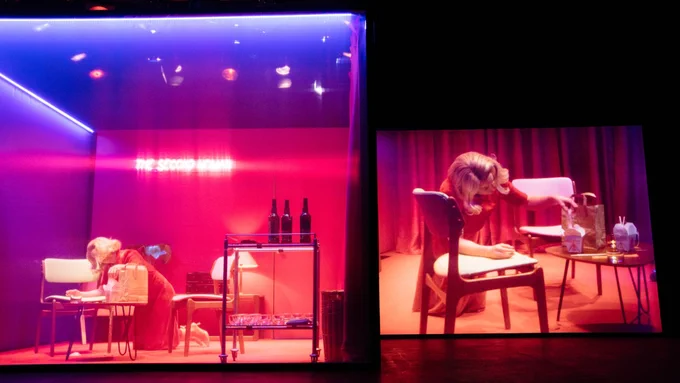 A person stands on their knees while reaching for the noodles on the table next to them. To the right, a screen projects the same scene from a different angle.
