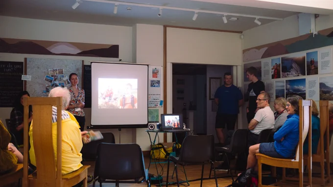 Faye Morrissey, a young woman in a floral shirt and black trousers, with a blue Armitt lanyard round her neck, stands to the left of a projector screen set up in The Armitt's Running Int Fells exhibition. People sit in chairs set up around the screen, waiting for the event to begin. To the right, Harrison Ward and Alex Staniforth stand together in conversation