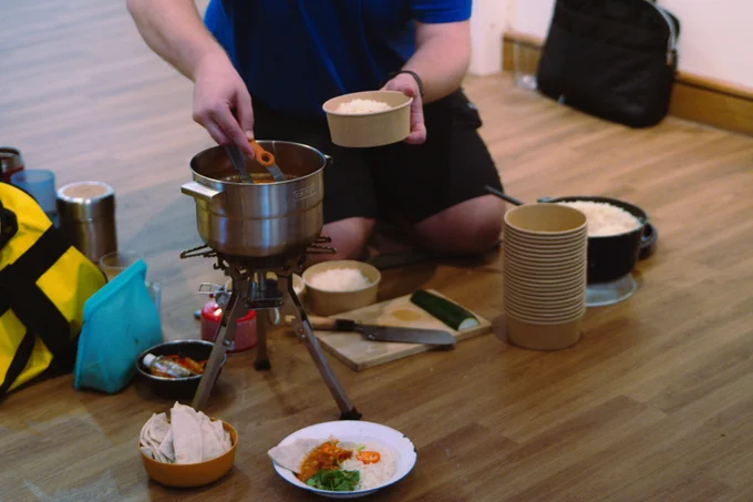 Harrison Ward, with his face out of frame, spoons vegan curry and white rice into cardboard food containers from a silver pan on a portable stove
