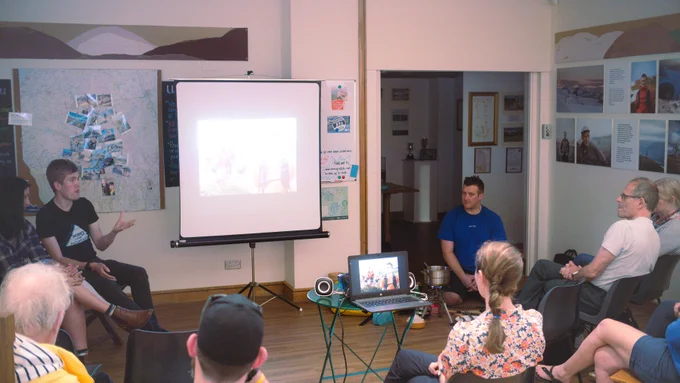 Alex Staniforth sits to the left of a projector screen in The Armitt gallery, with Harrison Ward on the right hand side with a portable stove and cooking gear set up around him. People sit on chairs facing Alex, Harrison and the screen, while Alex gestures mid-conversation