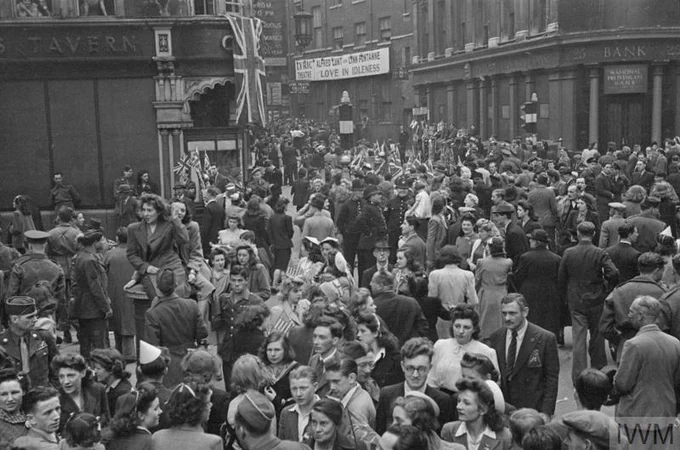 A crowd of people outside the gates of Buckingham Palace. In the left foreground, a soldier is holding a child aloft, perched on his right shoulder.