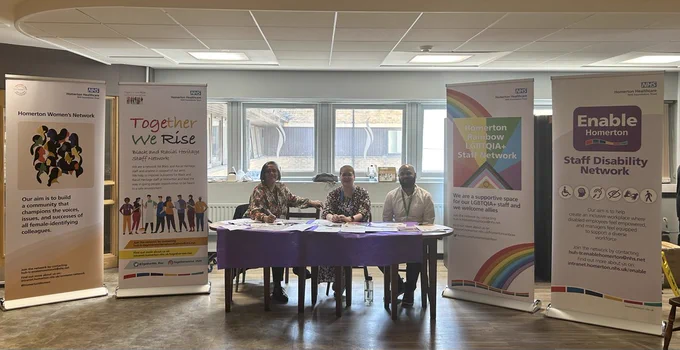Three network members are sitting behind a table, smiling at the camera. Either side of the table are pull up banners promoting the Women's Network, Together We Rise, Homerton Rainbow, and Enable Homerton