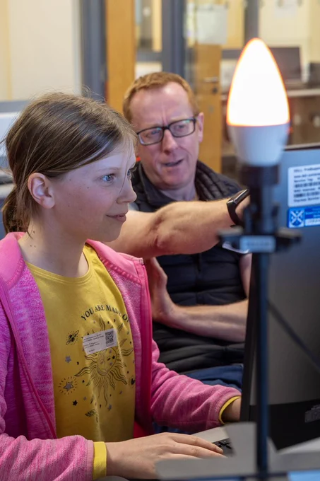 An attendee at the jam and their parent is shown on a computer with a lit up lightbulb in front of them.