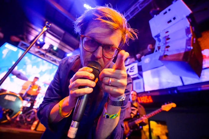 Close up of man holding a mic on stage, pointing at the camera.