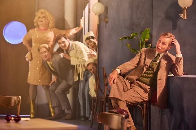 Malvolio in a brown suit leaning against the bar. Sailors, Toby Belch and Sir Andrew peer from behind a door at him. 