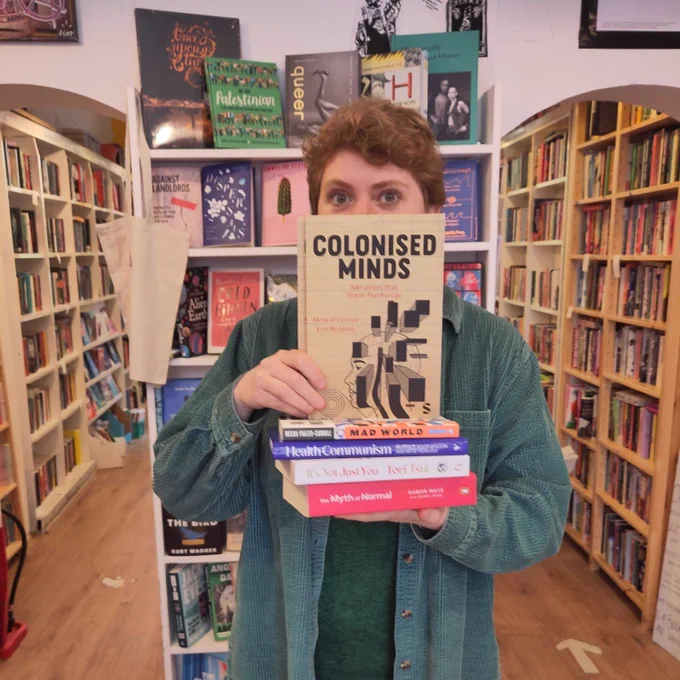 Owner of Lighthouse Mairi stands in the shop holding a stack of books with Colonised Minds face out on top. Only her eyes and nose are visible above the book. She's wearing a green corduroy shirt and has short ginger hair. 