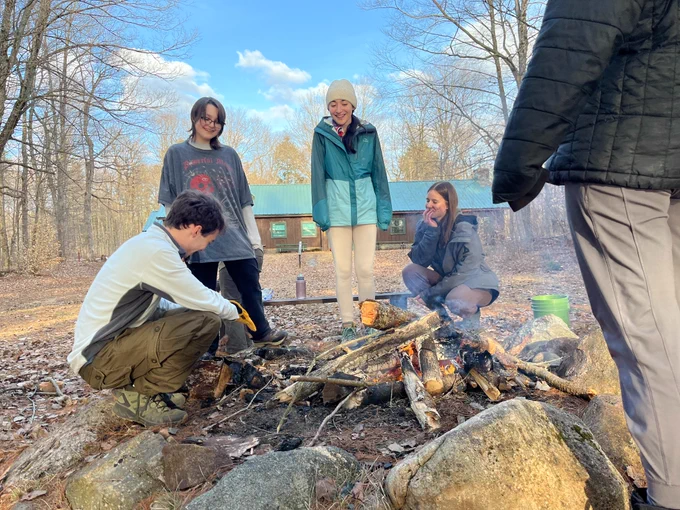 Five young people in a semicircle around a campfire during the day.
