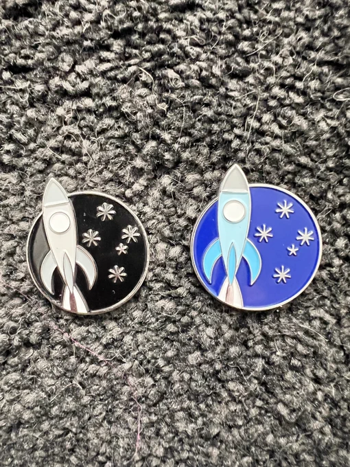 A pair of enamel pins/badges with the rocketship and stars logo from Continuum 15. The one on the left is black, while the one on the right is blue.