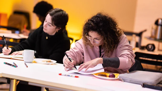 Two people sat at a table. They are both looking down, whilst they write and draw notes on pieces of paper and a notebook. Photograph by Paul Stringer. 