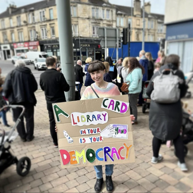 Kirsten holding a sign that reads ‘a library card in your hand is your democracy’ surrounded by a blurred-our crowd outside Cambuslang Library.