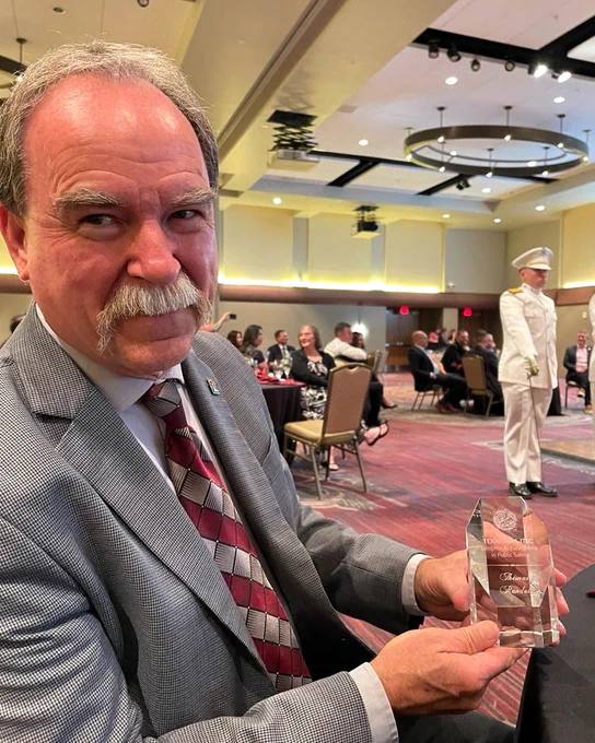 Tom Randall, senior public safety advisor, FirstNet Authority, smiles sitting in a conference room as he holds his lifetime achievement award in public safety from the Texas A&M University Internet2 Technology Evaluation Center