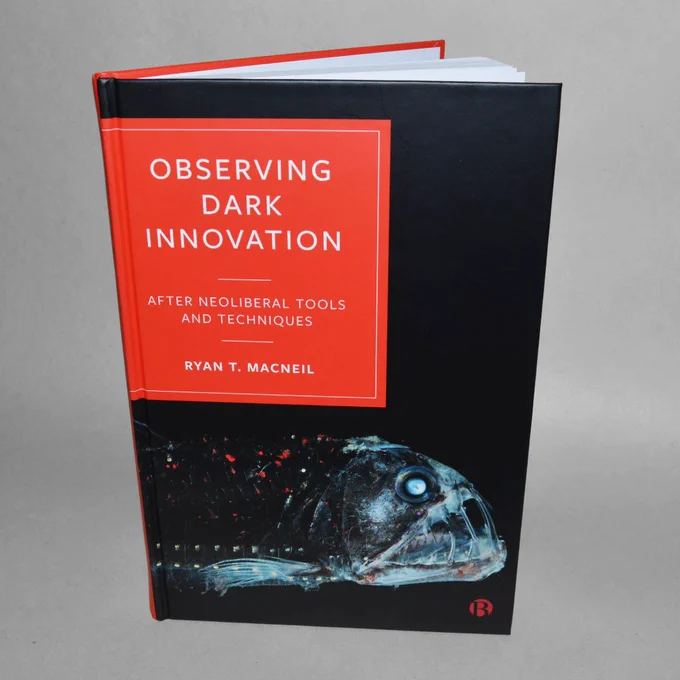 A photograph of the hard cover book, Observing Dark Innovation, by Ryan MacNeil. Cover features a black background with a photograph of a creepy deep sea fish with long translucent fangs. The book title and author name are imposed on a red square that matches the colour of spots of the body of the fish.