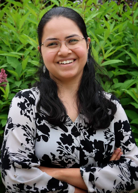 Dr. Aathmaja Anandhi Rangarajan smiles at the camera. Her arms are crossed, and she is standing in front of a flowering shrub. She is wearing a black and white floral blouse. 
