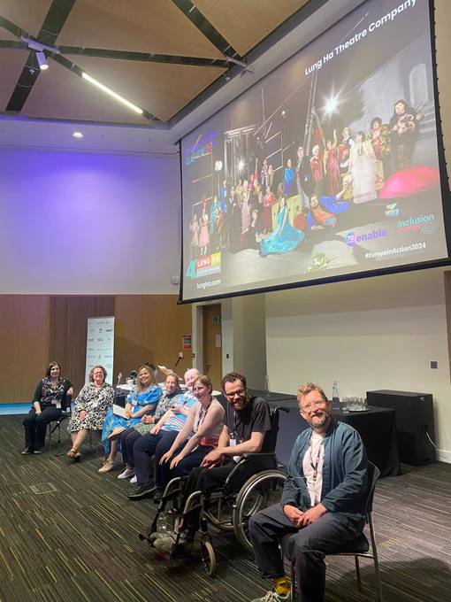 A line of people sittings on chairs and in wheelchairs on a stage, smiling at the camera. There is a screen with a colourful image projected behind them and above their heads. 