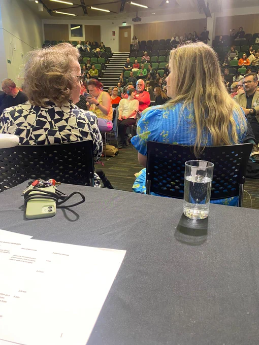 Picture taken from behind two people sitting in chairs, looking at each other. There is an audience in front of them. In the foreground there is a table covered in black tablecloth with a phone, white paper and a glass. 