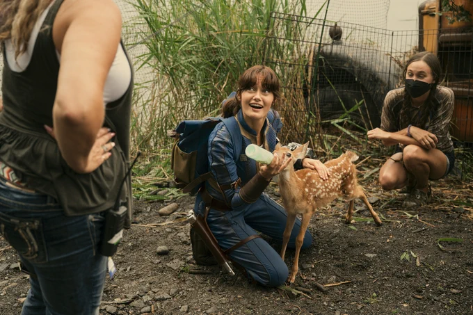 A behind-the-scenes shot on the Fallout set with Ella as Lucy kneeling down in the dirt in full Vault-Tec gear looking up and to the left at a crew member while feeding a fawn from a bottle with a wonderous smile on her face. 