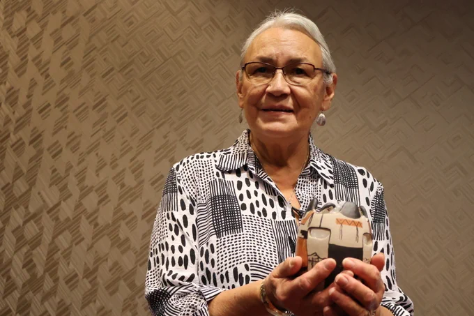 Honorable Sophie Pierre (Kootenay, Ktunaxa Nation) cradles a Tohono O'odham friendship pot in her hands and smiles in front of a textured brown wallpaper.