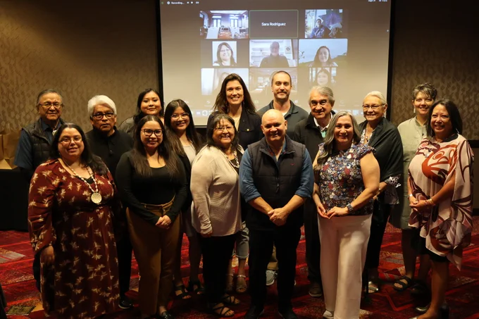 Group photo of NNI IAC and staff in front of a projector with faces of virtual meeting participants.
