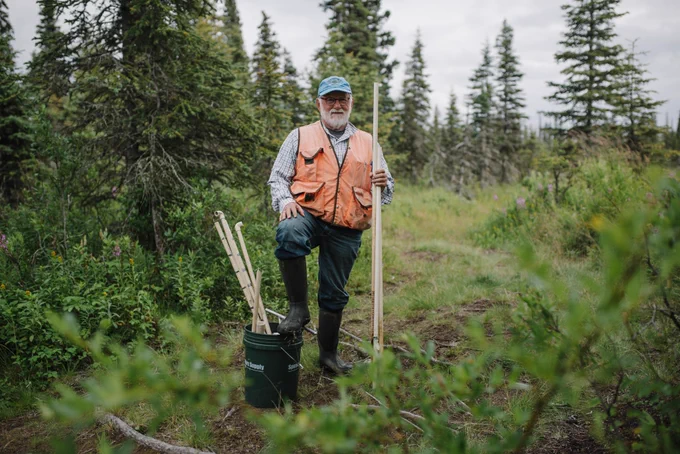 Dennis Whigham stands on a forest path lined with conifers, wearing an orange vest and a light blue cap.