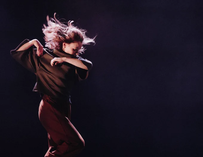 An image of a female dancer in a full body twist, her blonde hair flowing through the air, arms and elbows by her shoulders. She’s looking down to the stage floor wearing a dark olive top and brown trousers.