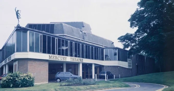Archive photo of the exterior of Mercury Theatre, a large 1970s building with a statue of Mercury on its root and the theatre name in bold letters above the entrance.