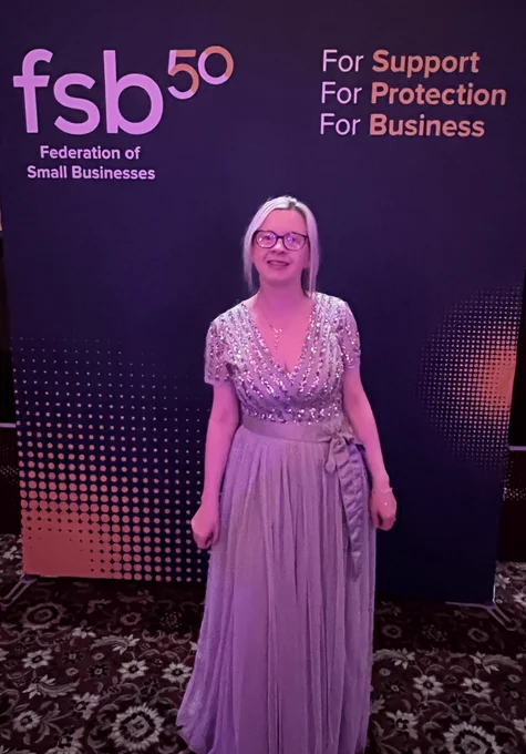 Hayley, a blonde white female wearing glasses and a very pretty sage green dress with green sparkly sequins all over the bodice, which has short sleeves and a v neck front, the skirt is full length and very floaty and has a sash tied around the middle in a bow. Hayley is stood in front of a Federation of Small Businesses board.
