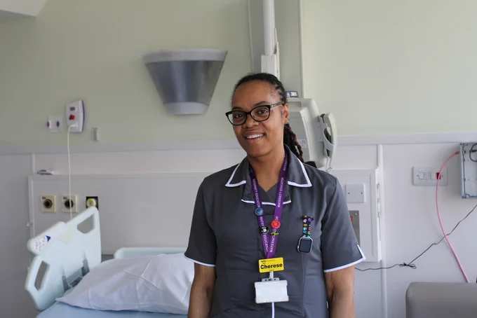 Cherese is wearing a grey dress with white piping, and she has a purple lanyard with a yellow name badge. She has her hair tied back and is wearing glasses. She is standing in a bay, with a hospital bed and equipment behind her.
