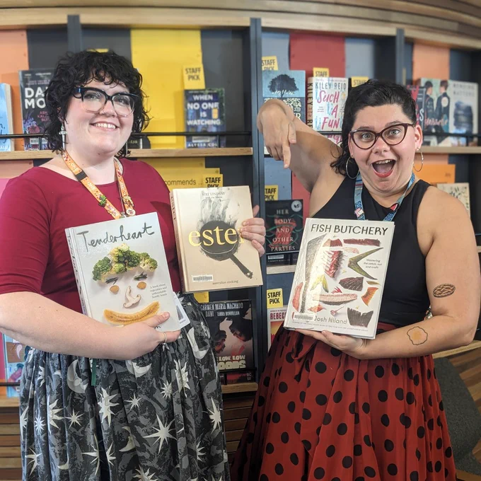 Library staff Amy and Connor holding copies of the books discussed in the post. Both are smiling in front of a book shelf in the library and Connor is pointing down at one of the books. 