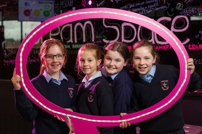 Four girls holding up a circular frame and smiling for a camera. Behind them there is reflective glass with illustrations and drawing on it. 