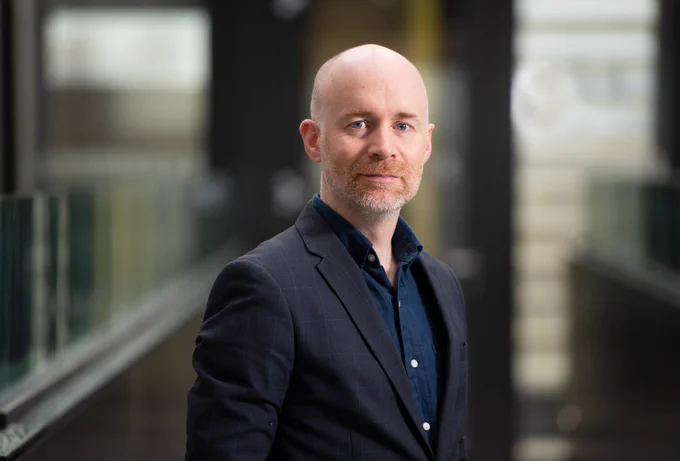 A photo of Professor Killian Hurley, who appears as a man with a bald head and a short beard. He is dressed in a professional manner, wearing a dark suit jacket over a navy shirt. He is standing in a modern academic setting, with architectural glass structures in the background. 
