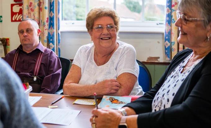 Image shows a group of people sitting around a table smiling and doing a craft activity.