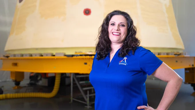 Lauren Fisher pictured standing in front of the launch vehicle stage adapter for NASA’s SLS (Space Launch System) rocket. Fisher is wearing a bright blue shirt with the Artemis logo on it. She has dark brown hair. Credit: NASA