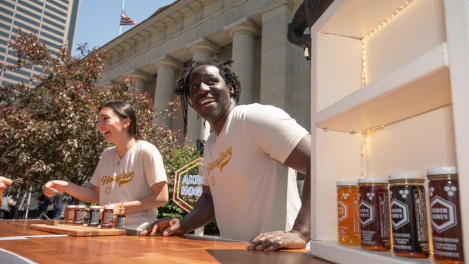 Brent Wesley, owner of Akron Honey, smiles at a table offering tastings of honey outside the Ohio Statehouse for Ohio Tourism Day in May 2023. Jars of different honey types are on display to the right. To the left is Wesley's helper interacting with one of the visitors.