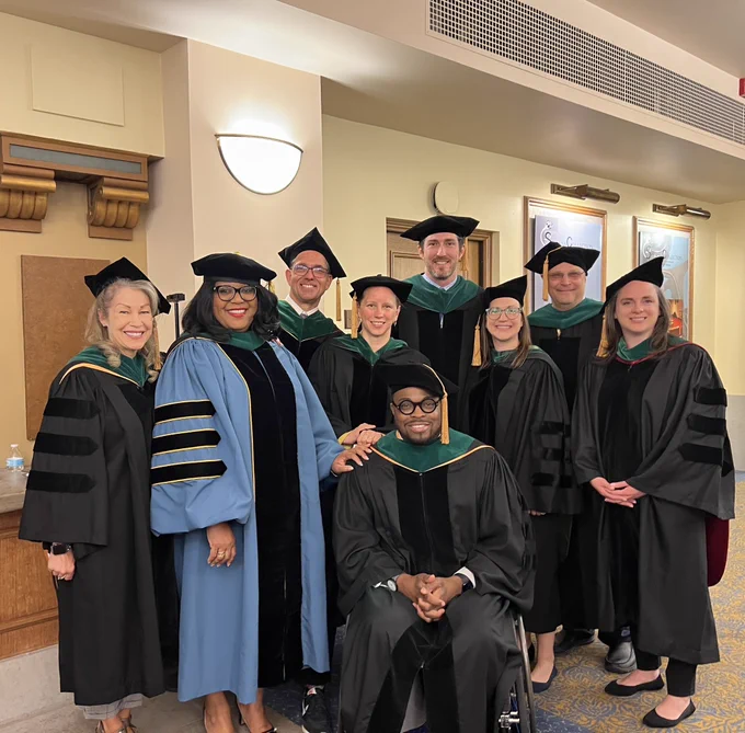 9 men and women in doctorate cap and gown snails for the camera. One is seated in a wheelchair.