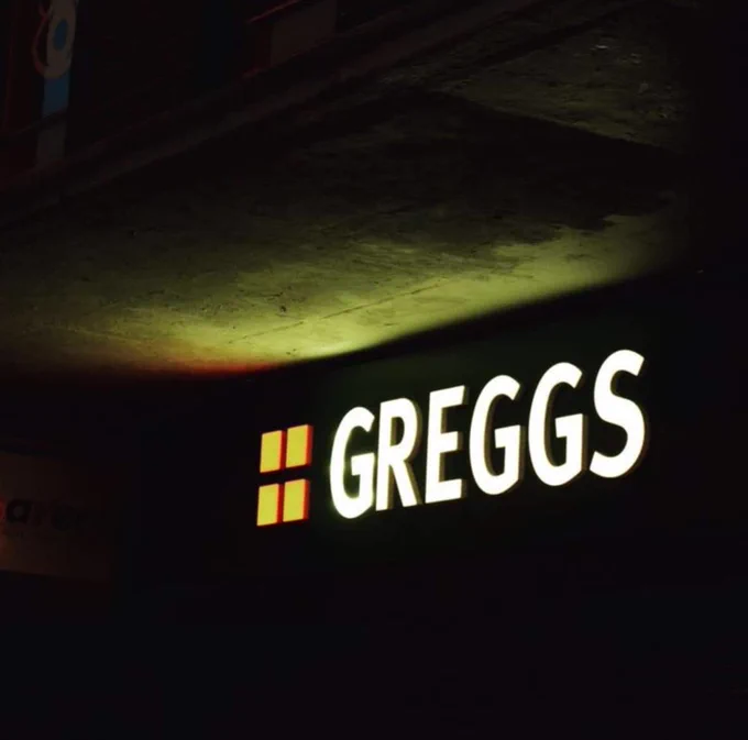 A Greggs sign lit up at night time, shining on the building above emulating the colours of The Northern Lights