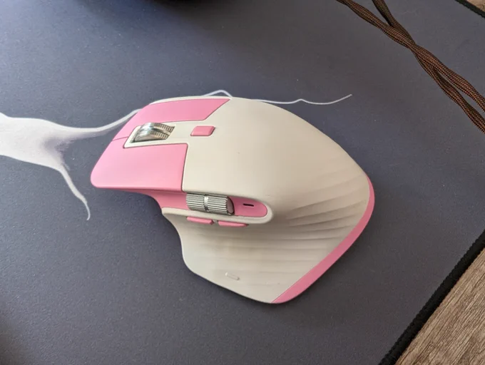 Logitech MX Master 3S mouse on a desk. The mouse is painted a custom pink and white colourway. The mouse is sitting on a grey and white desk mat.