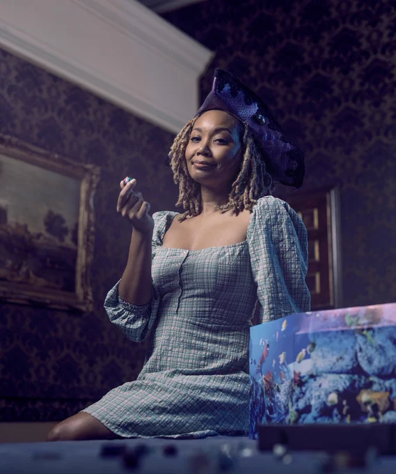 Photo of Aliza Pearl at Lumley Castle. She is half sitting on a table, looking into the camera, which is at a low angle on top of the table. She’s wearing a purple pirate hat with black lace trim, and holding up a D20. She is wearing a green and white gingham dress with puffed sleeves. Next to her on the table is her DM screen, which has a colorful underwater theme with fish and coral. On the table are lots of dice that are out of focus. She’s sitting in a an ornate, beautiful room at Lumley castle.