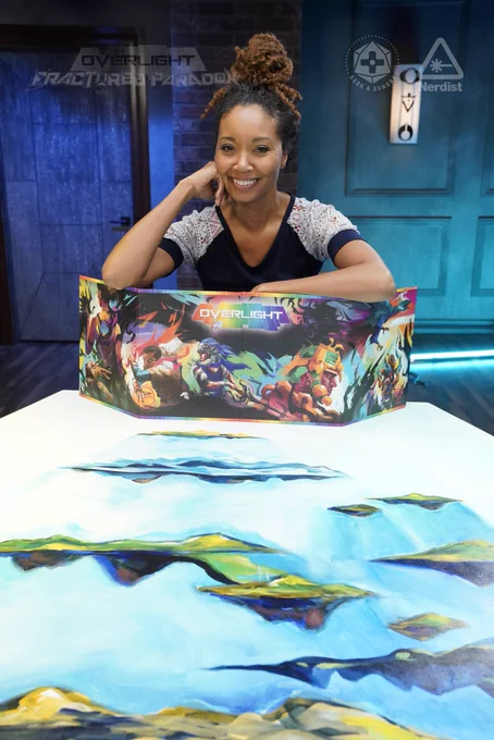 Photo of Aliza Pearl onset at Geek & Sundry. She is sitting behind a long table that has images of floating islands painted onto it. She leans over the top of a GM screen that says “Overlight” on it, and has beautiful, colorful art of characters from the Overlight game. She is smiling, and her face is leaning on one fist.
