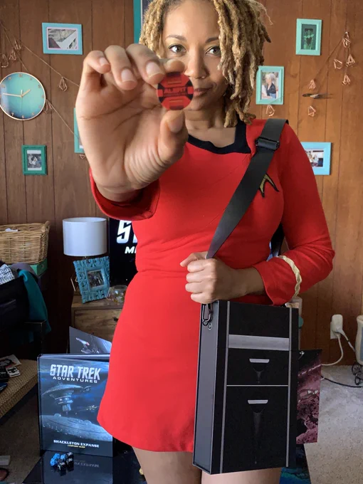 Picture of Aliza Pearl in a room with wood paneling and gray carpet. Behind her on the wood panel wall are turquoise framed photos and Star Trek adventures books and dice set up on a low coffee table. She is wearing a red Star Trek original series uniform dress, and holds the Modiphius Star Trek adventures Tricorder box slung over her shoulder. She is holding out a small red token that says “Alert” on it.