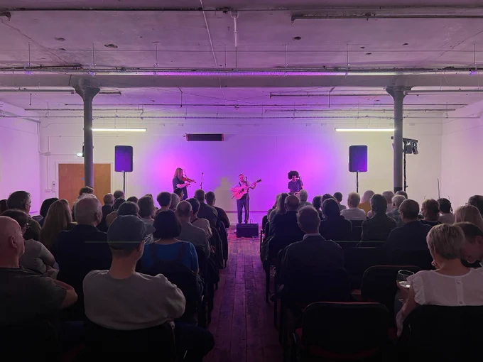 Photo from the back of a large room with white walls and purple stage lighting. Granny's Attic are playing onstage and there's an audience full of people listening.